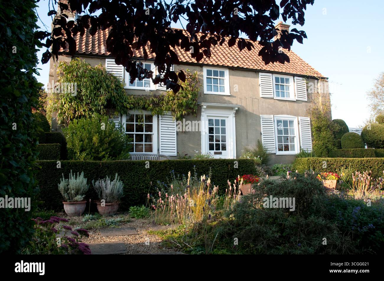 Un accogliente cottage con persiane bianche sorge in un giardino ben tenuto, caratterizzato da piante in fiore e siepi ben rifinite in un pomeriggio di sole. Foto Stock