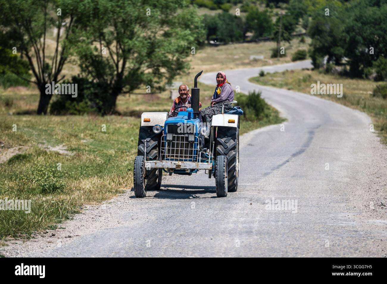 Un anziano agricoltore guida con sua moglie seduto sul parafango del loro vecchio trattore blu dai campi lungo una pista nella campagna turca Foto Stock
