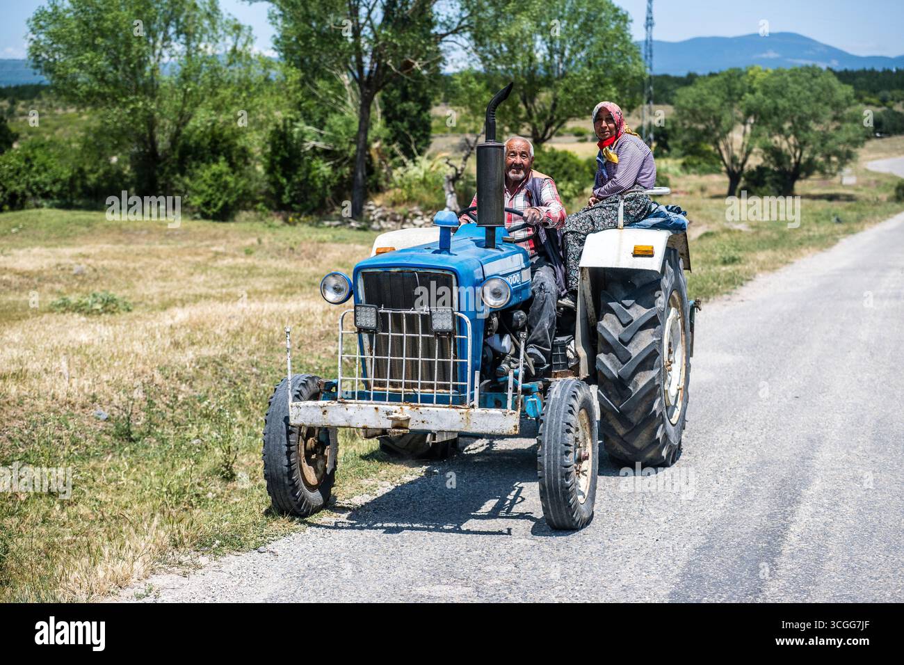 Un anziano agricoltore guida con sua moglie seduto sul parafango del loro vecchio trattore blu dai campi lungo una pista nella campagna turca Foto Stock