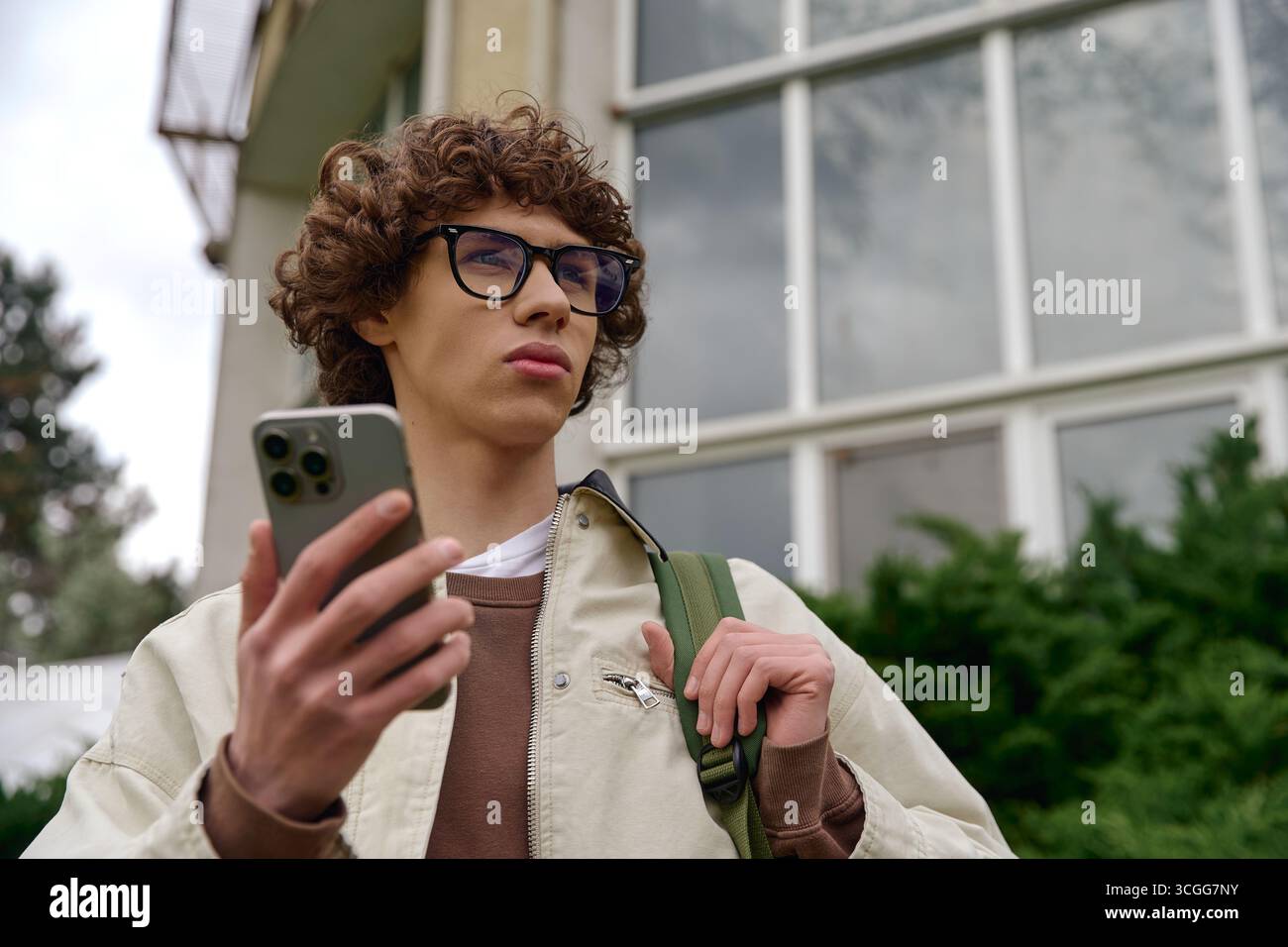 Un giovane con i capelli ricci cammina fuori, curiosamente controllando il suo smartphone. Foto Stock
