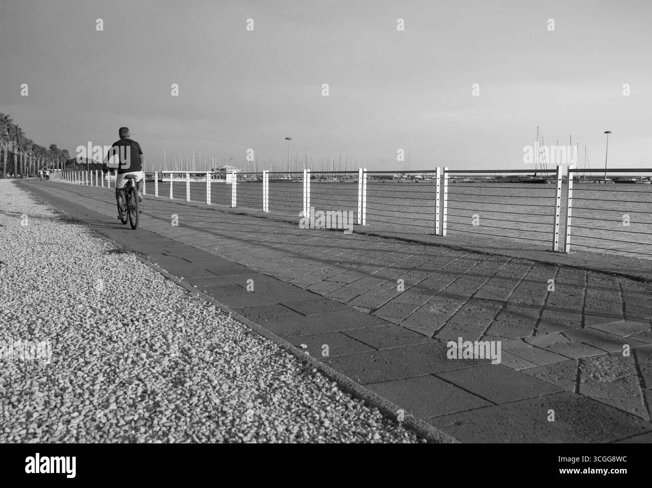 Un ciclista solitario pedala lungo una tranquilla passerella, con una pittoresca vista sul mare e barche in lontananza, catturate in bianco e nero. Foto Stock