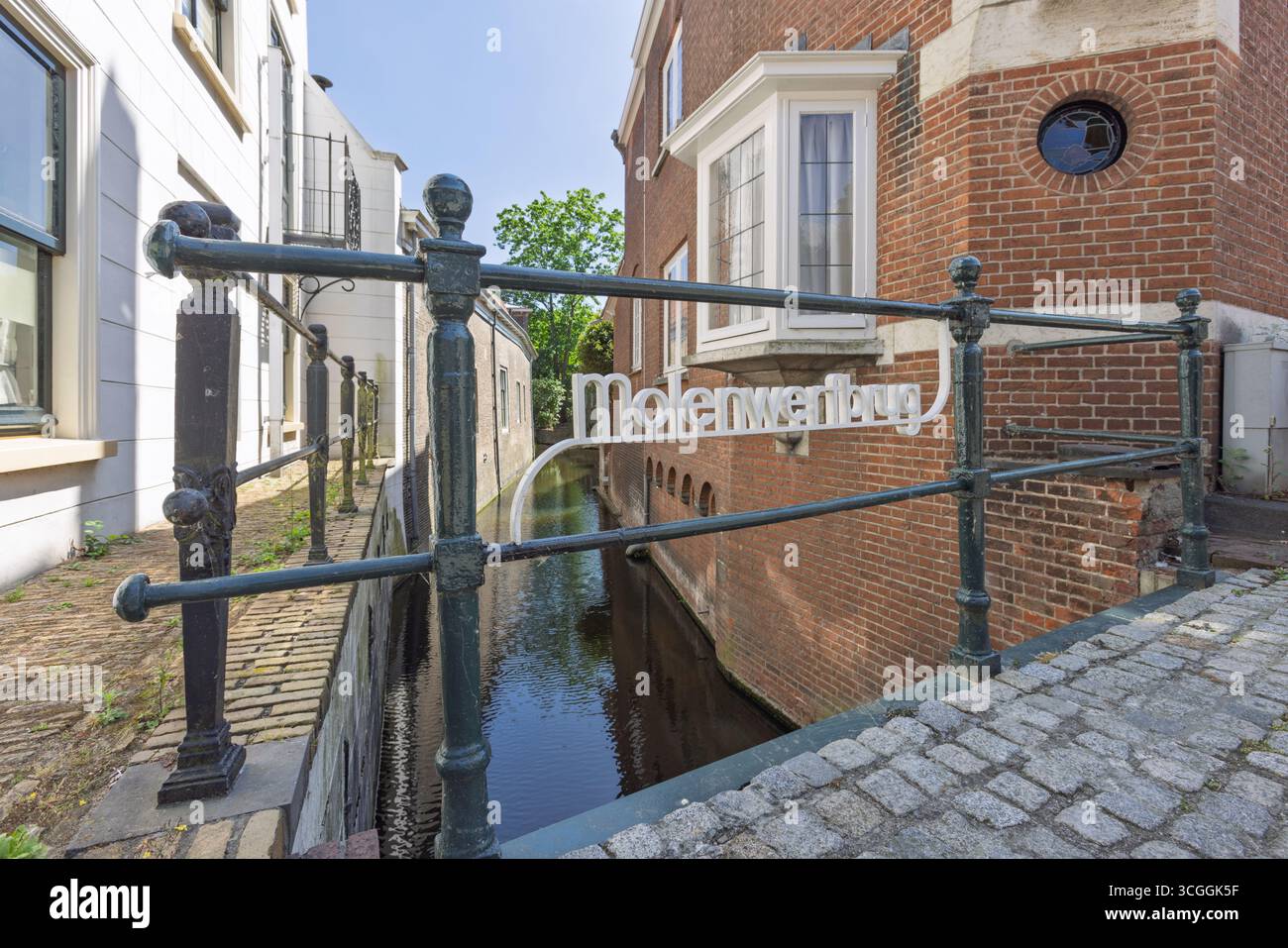Un affascinante ponte in pietra con una ringhiera decorativa e un cartello "Molenwerfbrug" che si estende su uno stretto canale della città di Gouda, circondato da una storica costruzione in mattoni Foto Stock