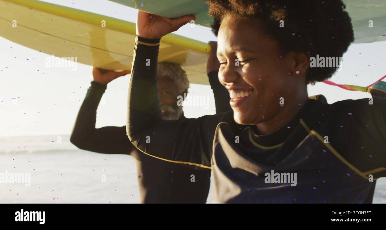 Donna e uomo afroamericani che camminano trasportando tavole da surf sopra la riva, con muta nera Foto Stock