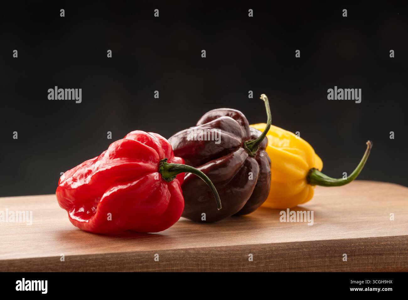 Tre peperoni Habanero freschi nei colori rosso, marrone cioccolato e giallo disposti su una tavola da cucina in legno su sfondo scuro Foto Stock