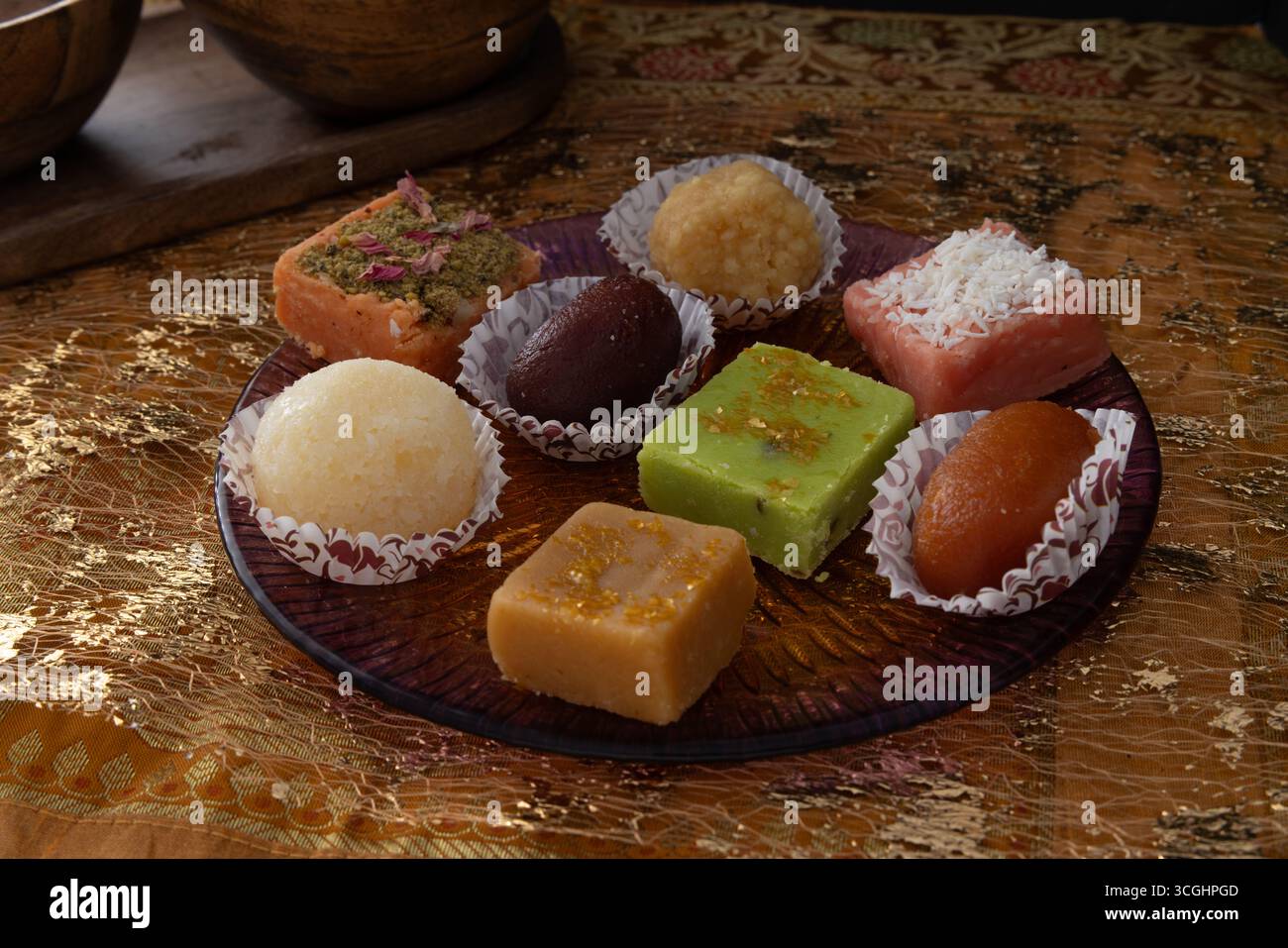 Dolci indiani assortiti disposti in tazze di carta su un piatto decorativo. Il tradizionale piatto mithai si stende su un panno a motivi geometrici per fotografare il cibo delle feste. 🔍 Foto Stock