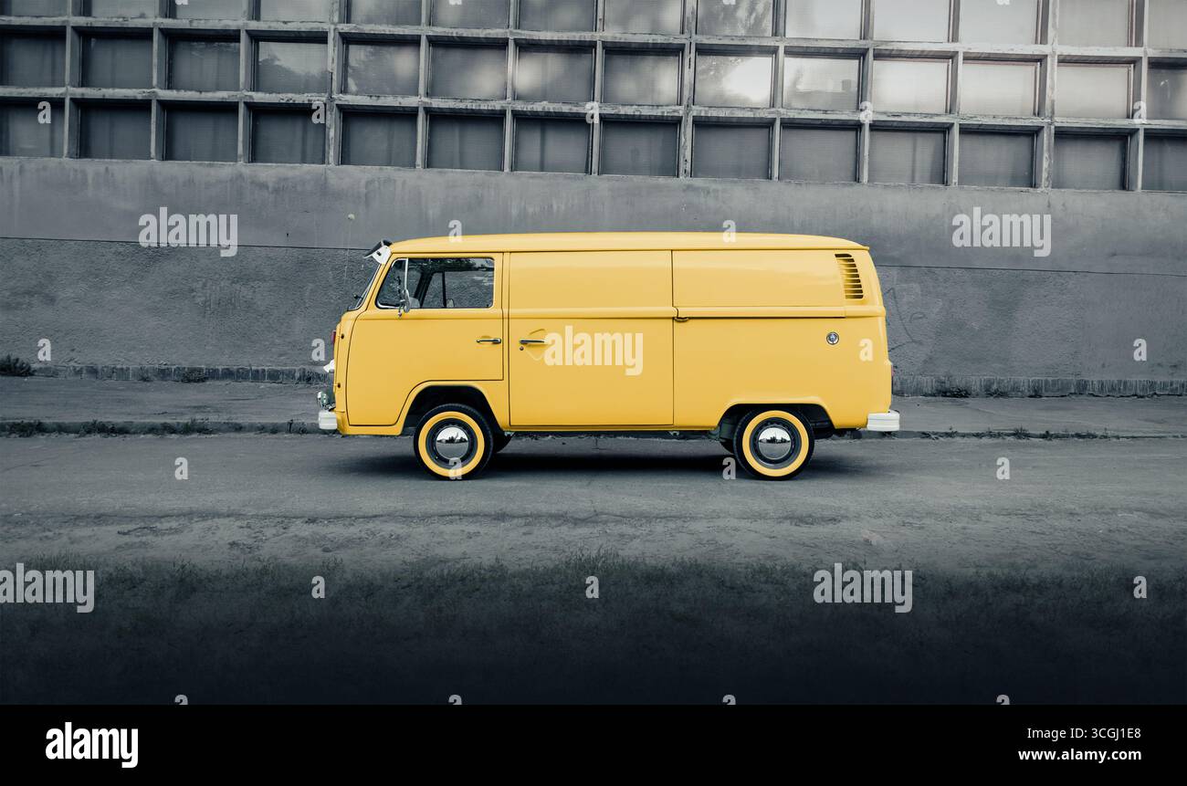 Pulmino giallo sulla strada di fronte a un muro di cemento grigio. Vista profilo laterale del furgone Volkswagen Transporter degli anni '1970 parcheggiato vicino a una parete con quadrati. Foto Stock