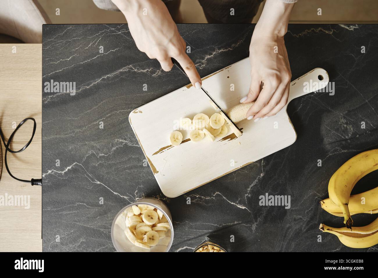 Una persona affetta banane fresche su un tagliere bianco in una cucina ben illuminata. Il piano di appoggio è dotato di una ciotola di banane e di un vaso di vetro. Esposizione alla luce solare Foto Stock