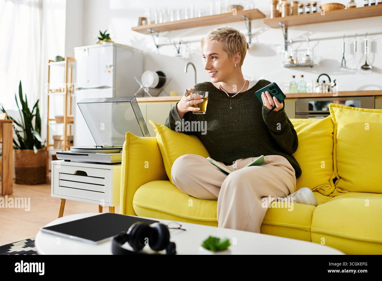Una giovane donna si rilassa su un divano giallo, bevi in mano, in una stanza luminosa e invitante. Foto Stock