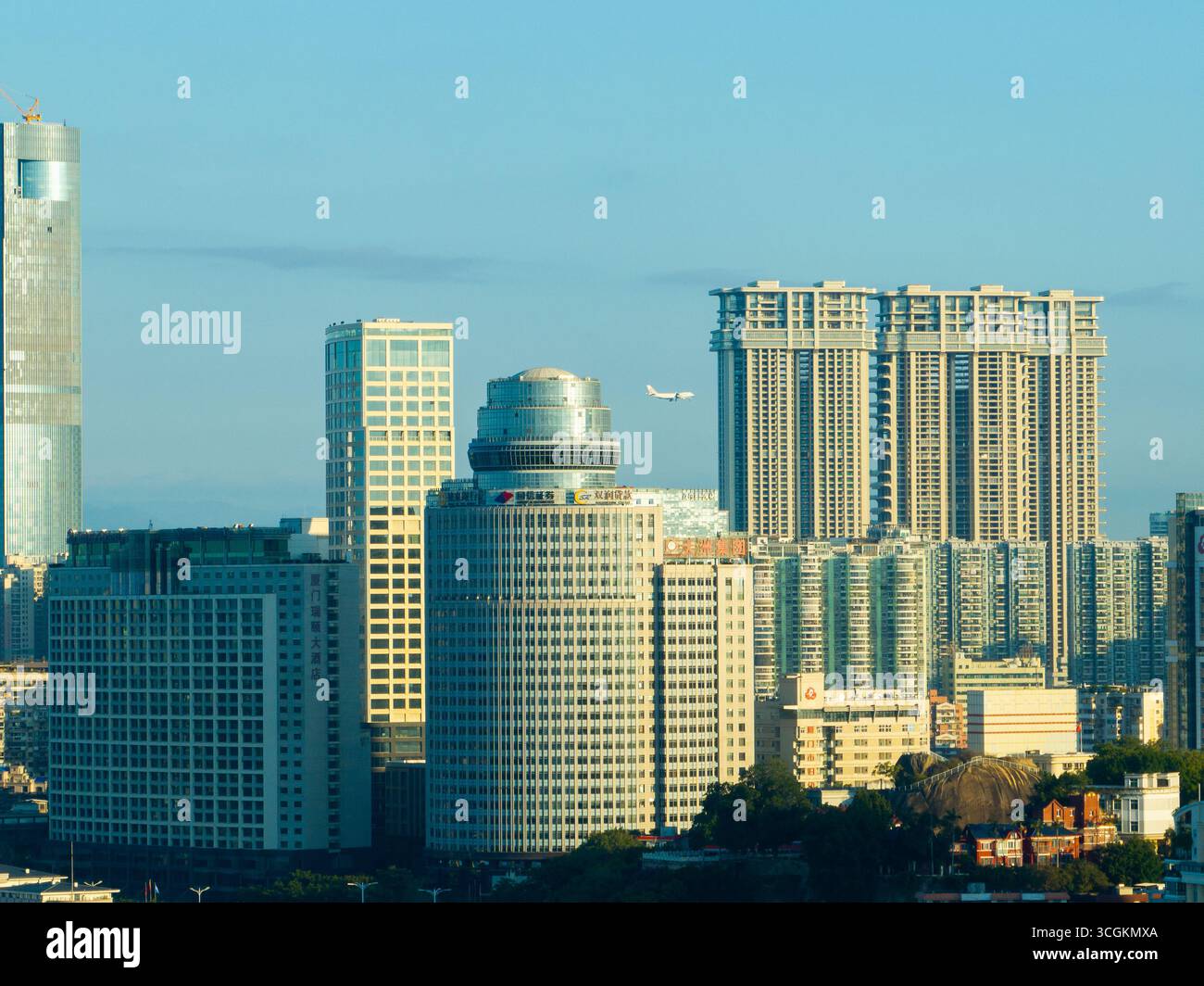 Il paesaggio urbano di Xiamen in Cina mostra molti alti edifici moderni e grattacieli prominenti che si avvicinano al cielo blu limpido mentre un aereo vola. Foto Stock