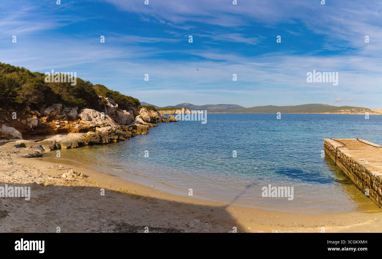 Tranquilla insenatura di Cala Dragunara vicino ad Alghero, in Sardegna, con acque turchesi cristalline, scogliere rocciose, sabbia dorata e un piccolo molo di pietra sotto un cielo blu. Foto Stock