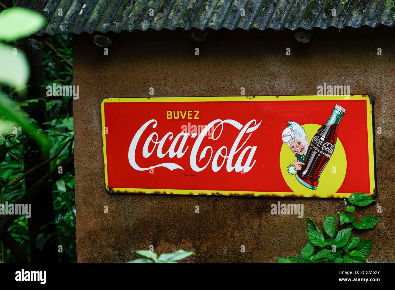 Insegna vintage Coca-Cola con testo francese su un muro rustico circondato dal verde. Foto Stock