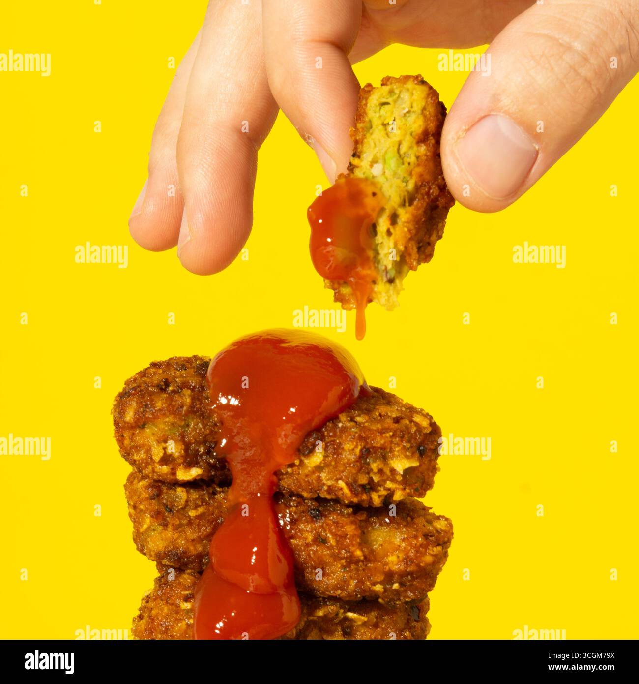 Mano che raccoglie un nugget croccante vegano da un mucchio su uno sfondo giallo, con una spruzzata di ketchup che cola dal nuggetto in alto Foto Stock