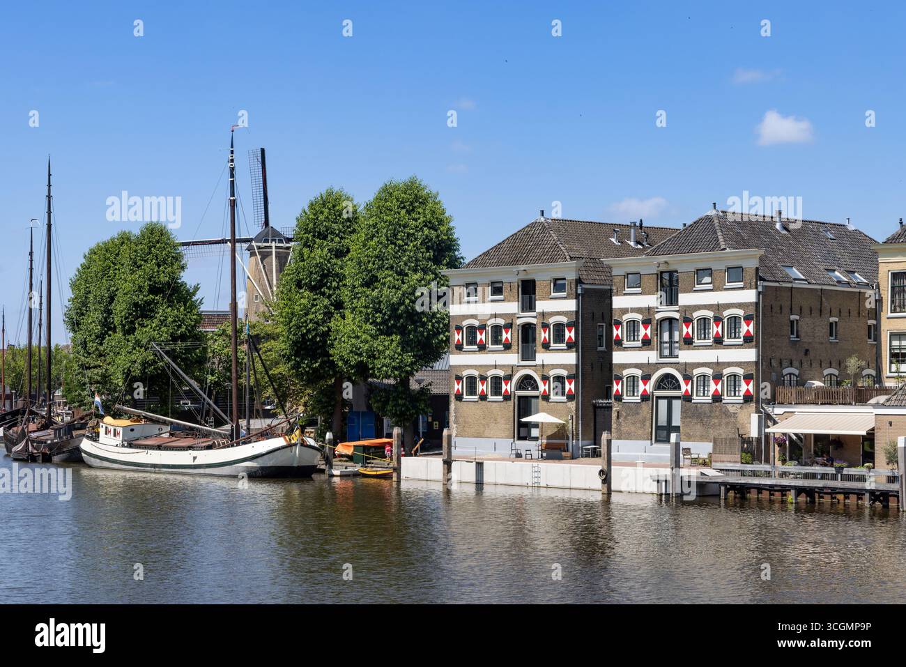 Una vista panoramica delle storiche case sui canali olandesi e delle barche a vela che costeggiano un ampio canale a Gouda, con un lontano mulino a vento visibile. Foto Stock
