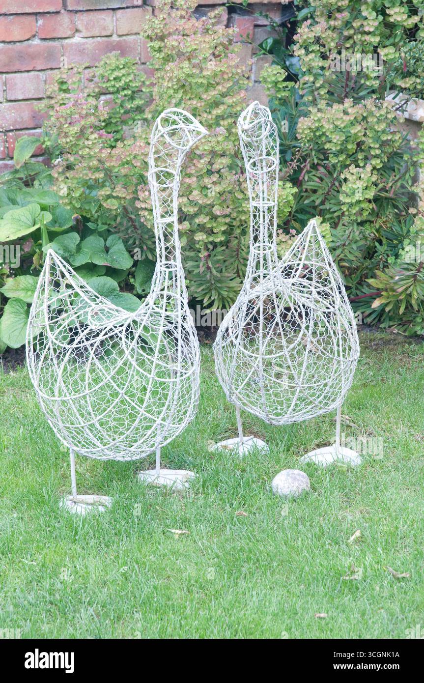 Due intricate sculture di oche metalliche sorgono sull'erba circondate da piante lussureggianti e pietre in un giardino luminoso. Foto Stock