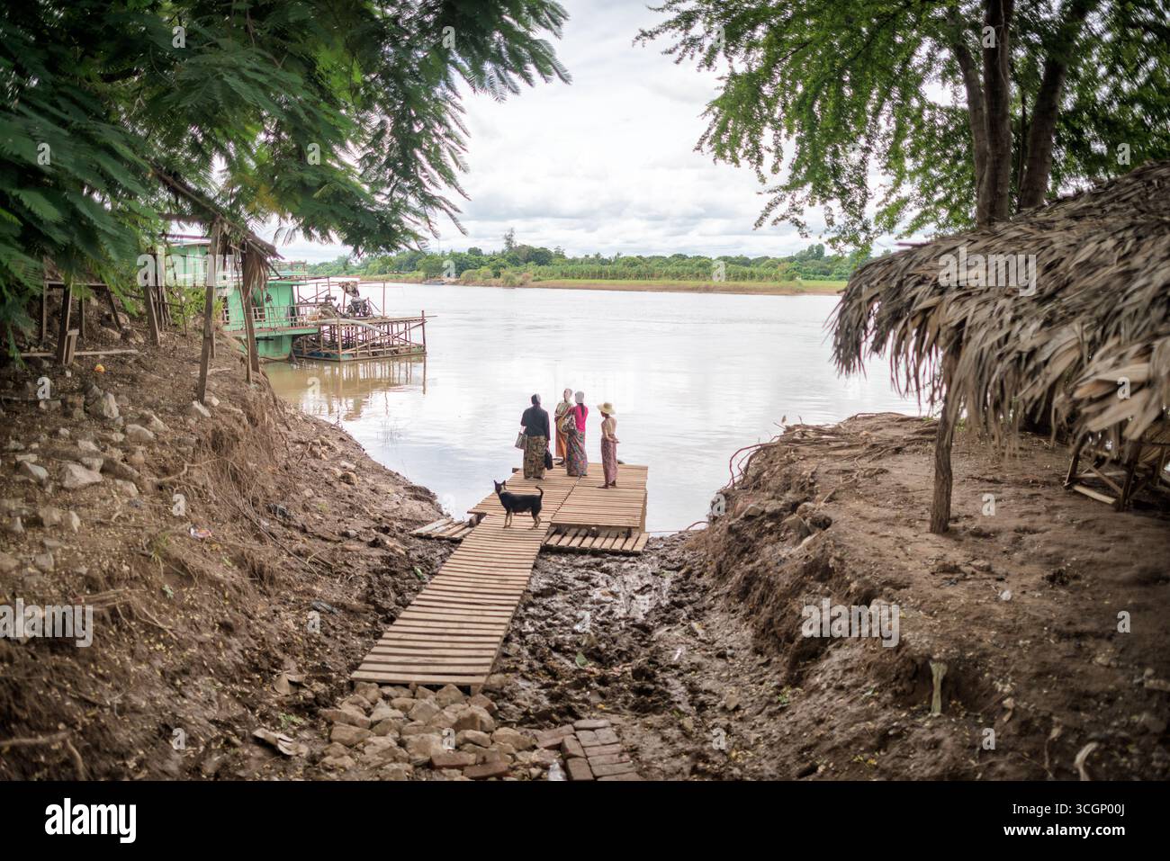 Le donne del fiume Myitnge aspettano il traghetto sul fiume presso il molo di legno Amarapura Myanmar // AMARAPURA, Myanmar (Birmania) - le donne e un cane attendono su un molo di legno per un traghetto sul fiume per attraversare il fiume Myitnge. Il fiume Myitnge è un importante affluente del fiume Ayeyarwady (noto anche come Irrawaddy River), cruciale per il trasporto locale e il commercio nel Myanmar centrale. Questa traversata in traghetto si trova vicino ad Amarapura, una storica ex capitale nella regione di Mandalay. I traghetti e le barche fluviali sono essenziali per gli spostamenti quotidiani e il commercio attraverso le ampie reti fluviali del Myanmar, collegando le comunità. Ama Foto Stock