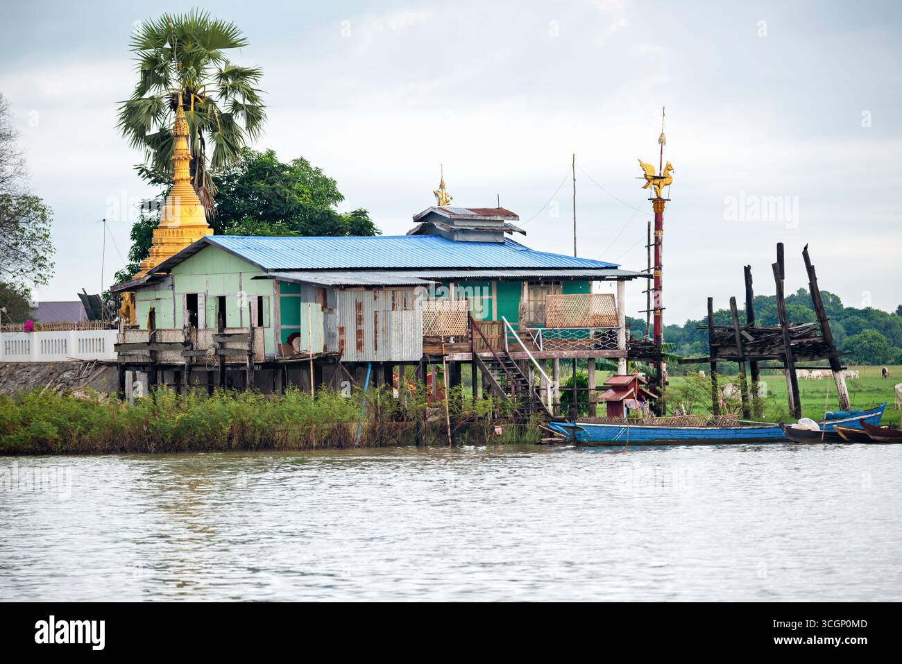 Ayeyarwaddy River Stilt House con Golden Pagoda e Hintha Bird Mandalay Myanmar // MANDALAY, Myanmar — Una tradizionale palafitta si trova sulle rive del fiume Ayeyarwady (noto anche come Irrawaddy River o Ayeyarwady River) a Mandalay. La scena presenta una pagoda dorata (stupa) e un'importante scultura dorata dell'uccello Hintha (noto anche come uccello Hamsa), che è un simbolo significativo nella cultura birmana, spesso rappresentando la purezza e la prosperità. Le palafitte si trovano comunemente lungo il fiume Ayeyarwady, fornendo case per le comunità adattate ai livelli fluttuanti dell'acqua del fiume. Questi Foto Stock