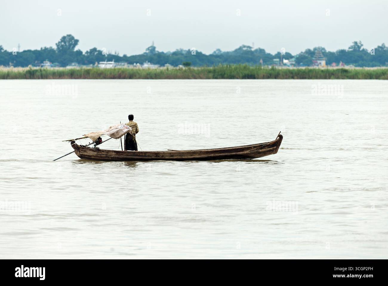 Fiume Ayeyarwady tradizionale battello fluviale con persona Mandalay Myanmar // MANDALAY, Myanmar — Una tradizionale barca fluviale in legno con una persona a bordo naviga sul fiume Ayeyarwady (noto anche come Irrawaddy River o Ayeyarwady River). Queste imbarcazioni lunghe e strette sono comunemente utilizzate per il trasporto locale, la pesca e il commercio. L'Ayeyarwady è la via d'acqua più lunga e vitale del Myanmar, che si estende per circa 2.170 chilometri (1.350 miglia). È una caratteristica centrale della vita a Mandalay, la seconda città più grande della nazione. Questa ex capitale reale si affida al fiume per la sua economia e il suo patrimonio culturale. Foto Stock
