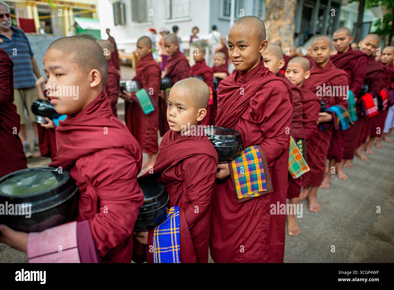 Monastero di Mahagandayon buddista Novices Alms Procession Mandalay Myanmar // MANDALAY, Myanmar — i novizi buddisti partecipano alla processione quotidiana delle elemosine (Pindapata) presso il monastero di Mahagandayon ad Amarapura, una cittadina di Mandalay. Questo prestigioso college monastico è uno dei più grandi del Myanmar, che ospita migliaia di monaci e novizi. Ogni mattina, si allineano per ricevere offerte di cibo dai devoti, una pratica fondamentale nel Buddhismo Theravada. Questo rituale si verifica prima del loro pasto principale della giornata. La processione è un evento culturale e religioso significativo, che riflette la profonda devozione della loca Foto Stock