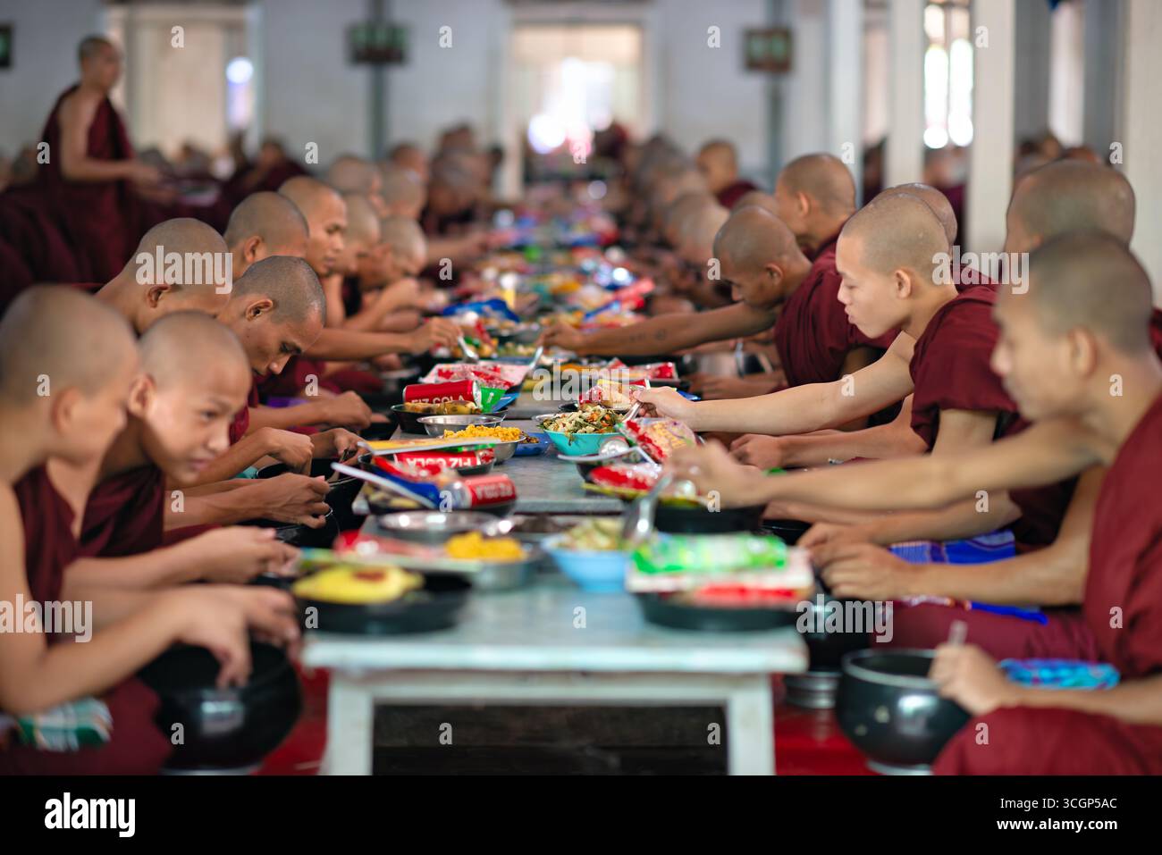 Mahagandayon Monastery Dining Hall Novices Lunch Mandalay Myanmar // MANDALAY, Myanmar - i novizi si siedono per il loro pasto a mezzogiorno nella sala da pranzo del Monastero di Mahagandayon (noto anche come Monastero di Mahagandayone) ad Amarapura, vicino a Mandalay. Essendo uno dei college monastici più grandi e rispettati del Myanmar, Mahagandayon è la sede di migliaia di monaci e novizi. Il pranzo quotidiano in comune è un aspetto fondamentale della loro vita monastica disciplinata, aderendo alle tradizioni buddiste, dove i pasti vengono consumati generalmente prima di mezzogiorno. Questo importante monastero funge da centro vitale per l'educazione buddista e Foto Stock