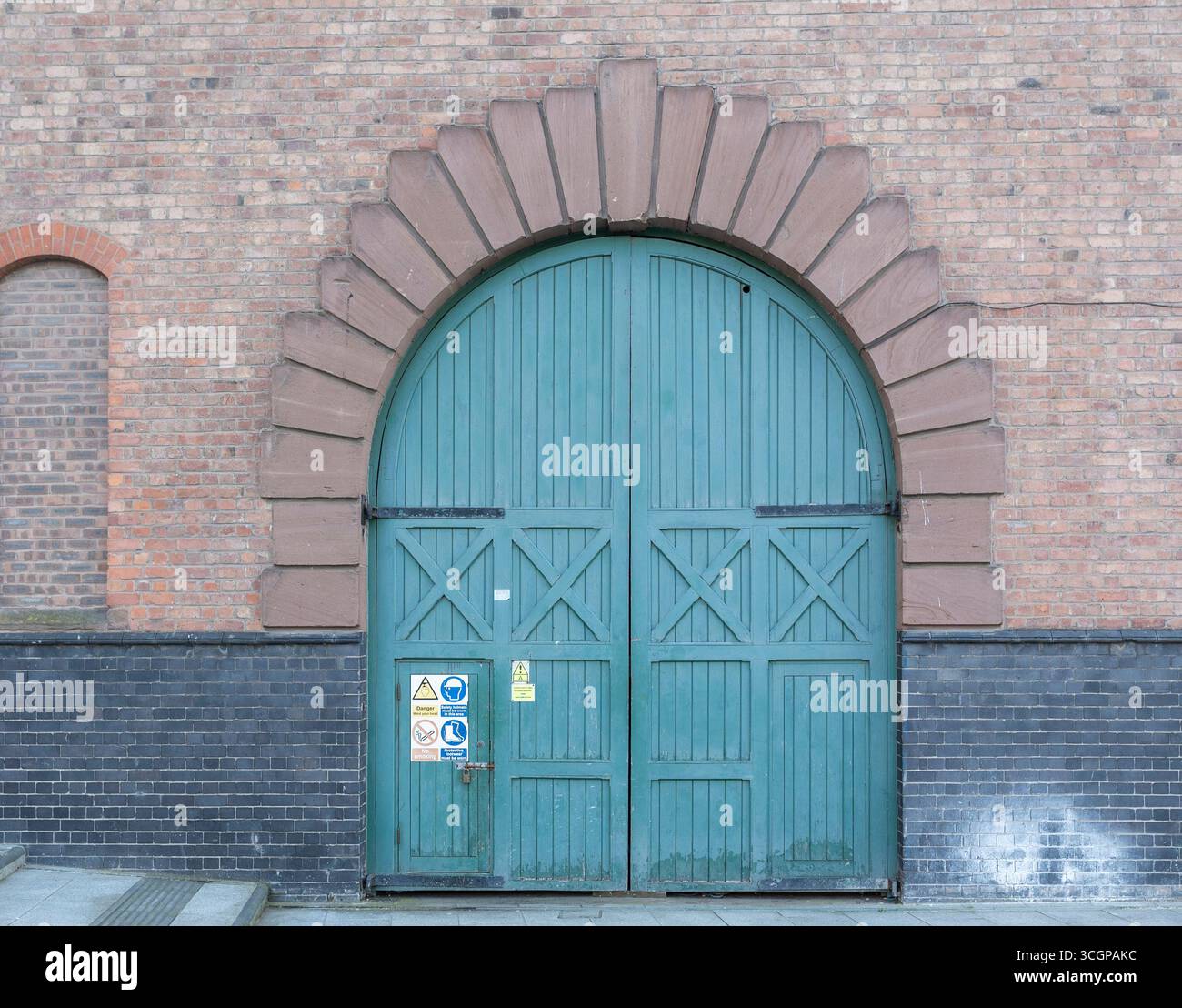 Una porta industriale in legno con un design ad arco verde adagiato su una facciata in mattoni, che mostra segnali di avvertimento ed elementi architettonici urbani. Foto Stock