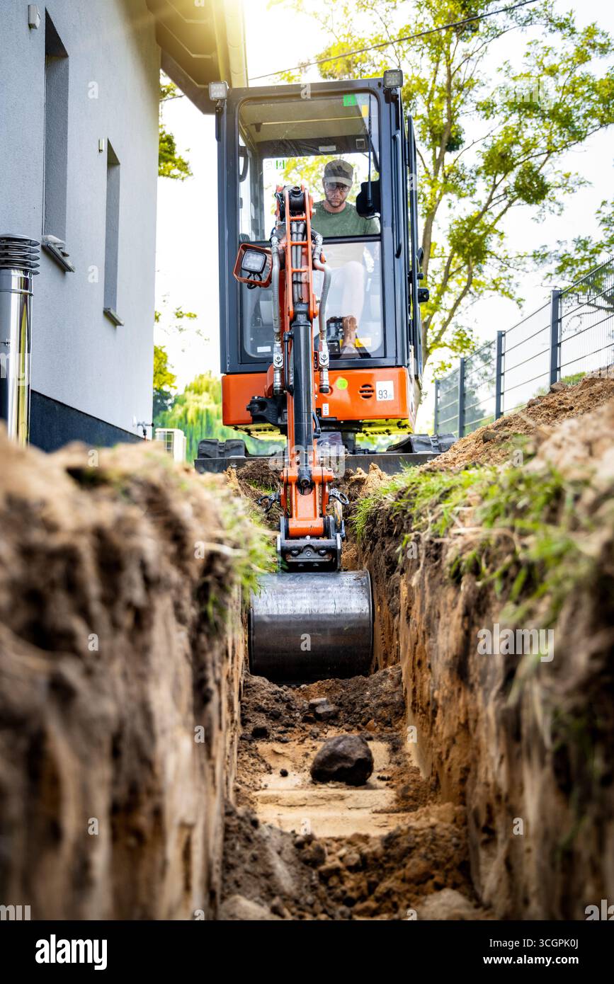 Miniescavatore scavando una trincea accanto a una casa Foto Stock