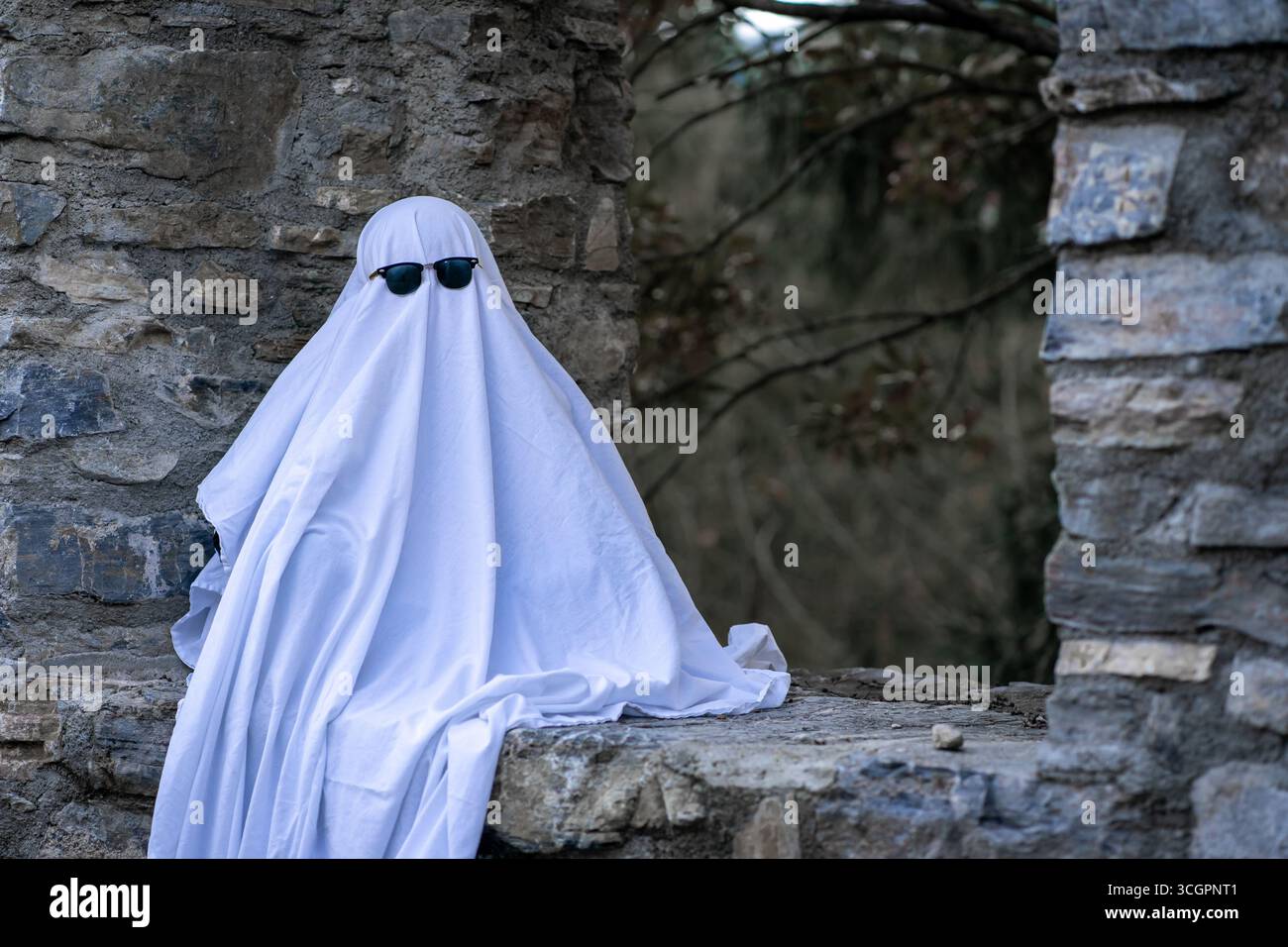 Un bambino, avvolto in un foglio bianco che scorre e indossa occhiali da sole, siede su un muro di pietra. Foto Stock