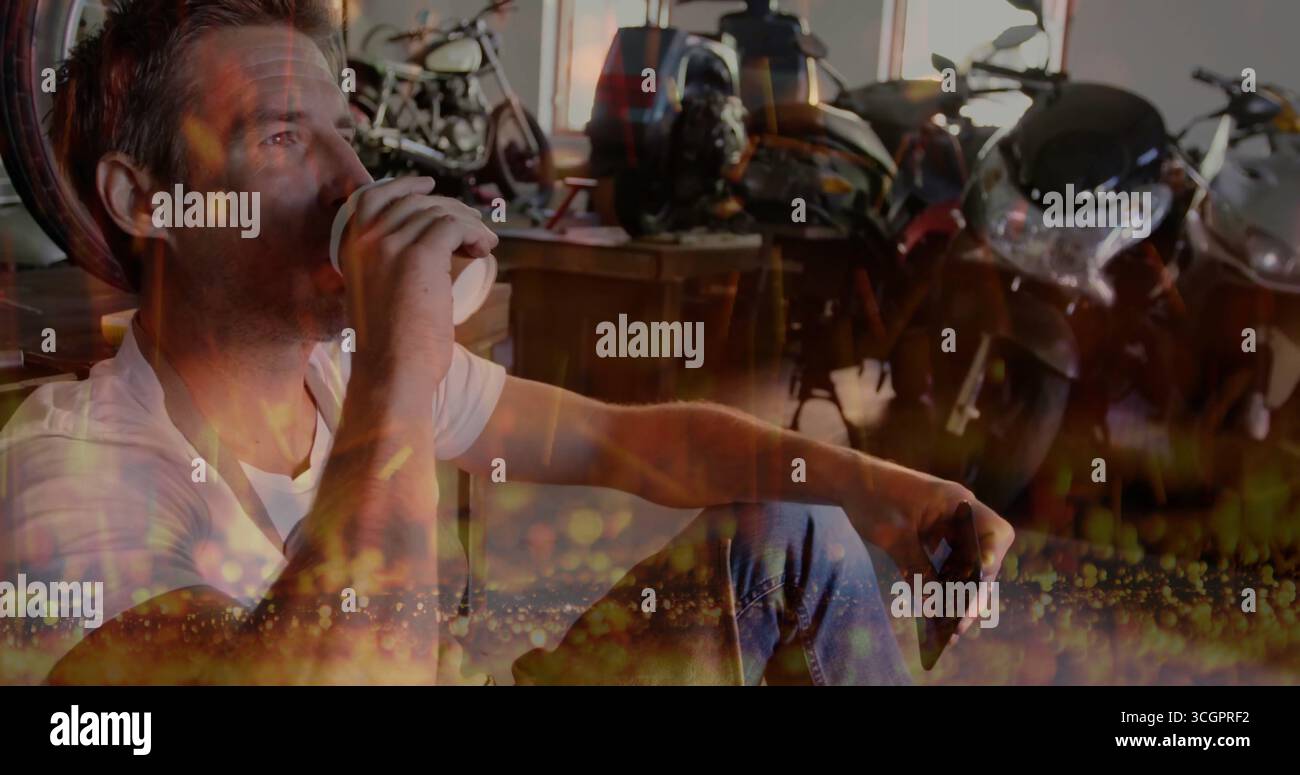 Uomo che tiene in mano una tazza di caffè e uno smartphone, appoggiato al banco di lavoro in garage con motociclette Foto Stock