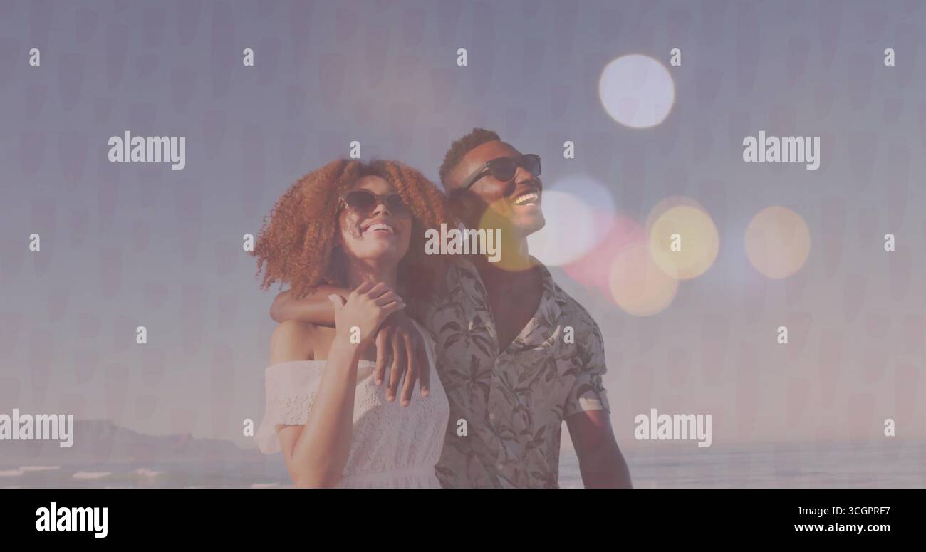 Coppia sorridente che indossa un abito a spalla, una camicia floreale e occhiali da sole in piedi sulla spiaggia in riva all'oceano Foto Stock