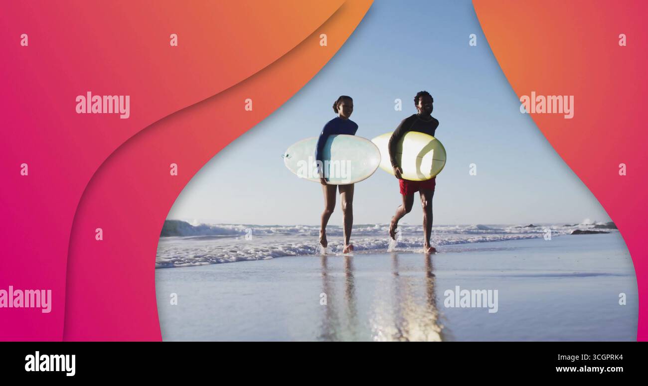 Coppia che cammina indossando muta che trasporta tavole da surf sulla spiaggia bagnata, con onde che si riflettono sulla sabbia Foto Stock