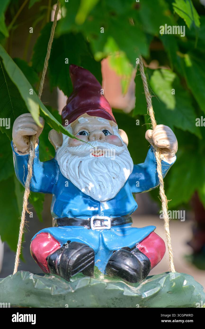 Uno gnomo colorato da giardino con un cappello rosso e una barba bianca oscilla da una corda sotto foglie verdi brillanti. Foto Stock