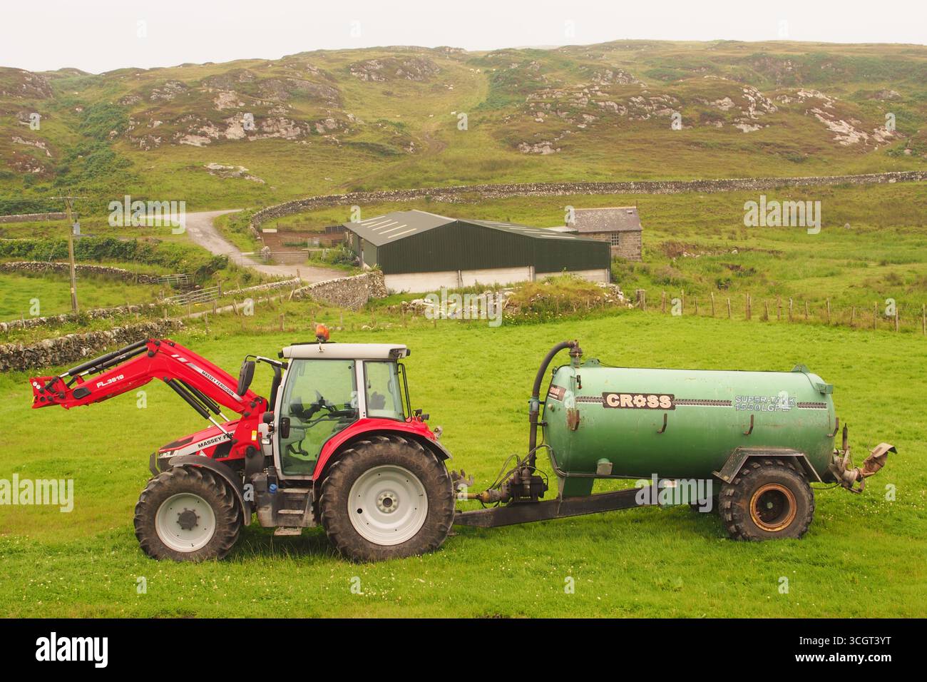 Un trattore Massey Ferguson, rosso e bianco, con rimorchio per concime liquido a croce verde, in un campo con un edificio agricolo sullo sfondo. Islay, Scozia Foto Stock