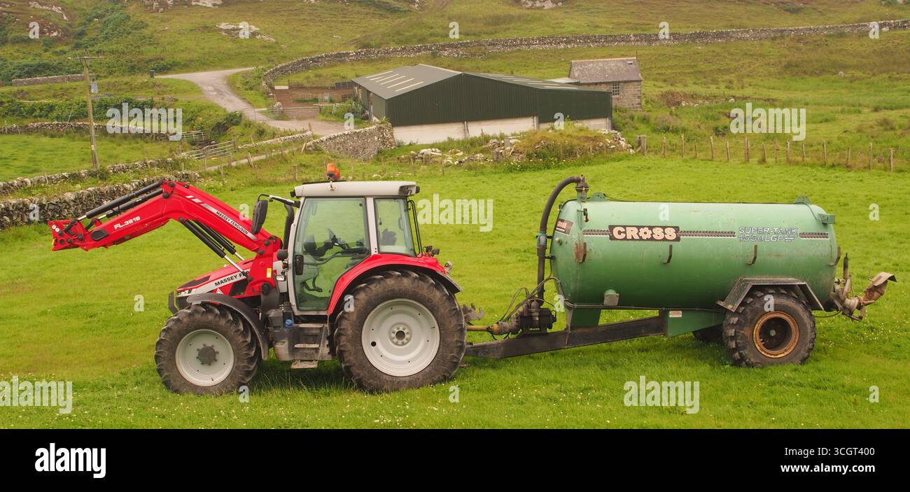 Un trattore Massey Ferguson, rosso e bianco, con rimorchio per concime liquido a croce verde, in un campo con un edificio agricolo sullo sfondo. Islay, Scozia Foto Stock
