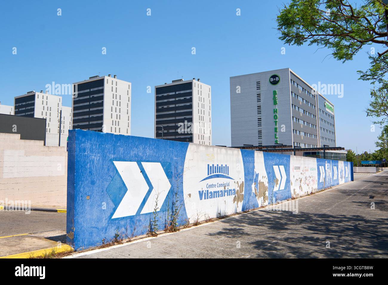 Viladecans. Barcellona - 29 agosto 2025: Una barriera in cemento dipinto con il logo del centro commerciale Vilamarina in primo piano, con il B B Hotel in Foto Stock