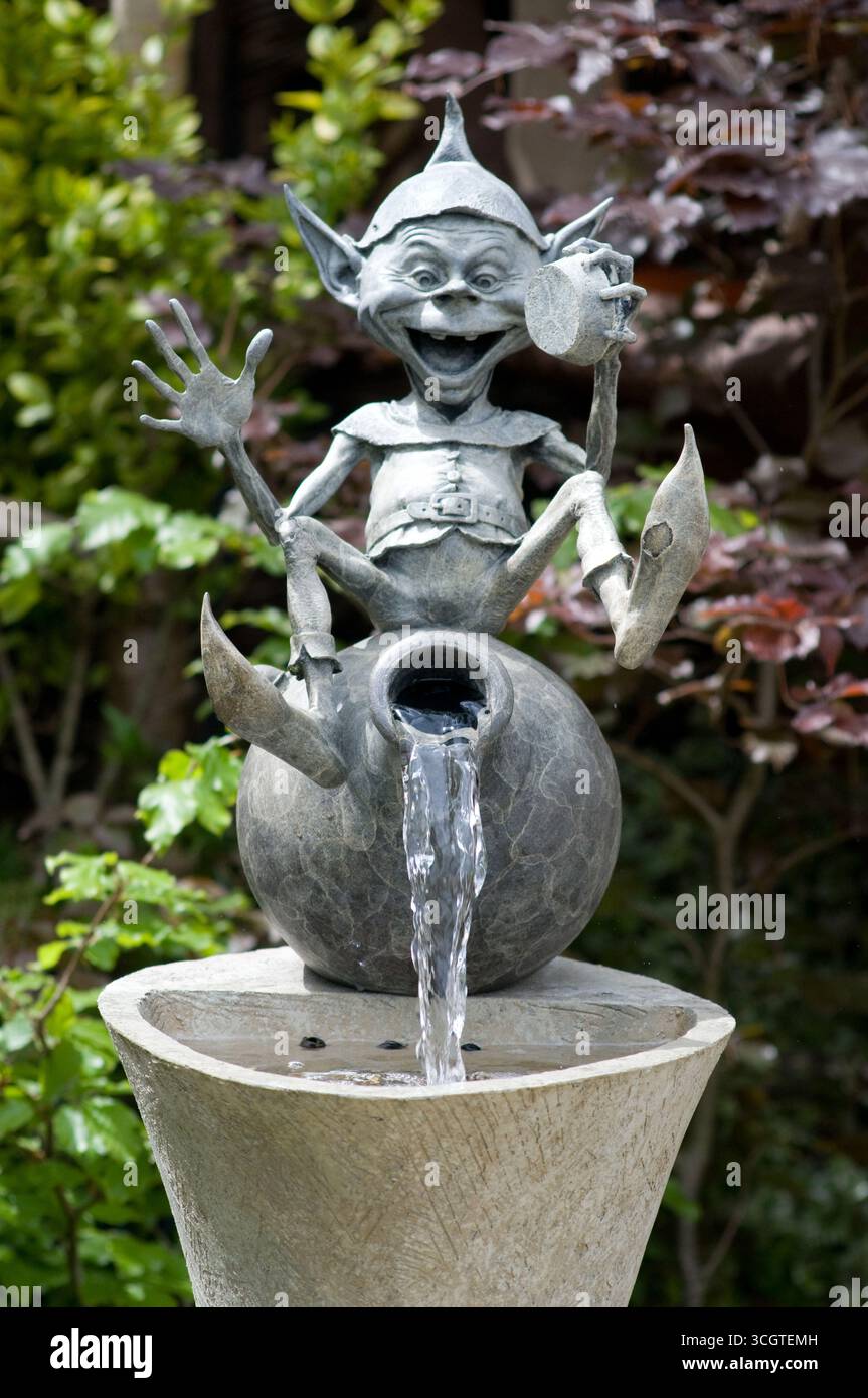 Una maliziosa statua di goblin siede in cima a un vaso rotondo, spruzzando acqua gioiosamente in un vivace giardino pieno di vegetazione. Foto Stock