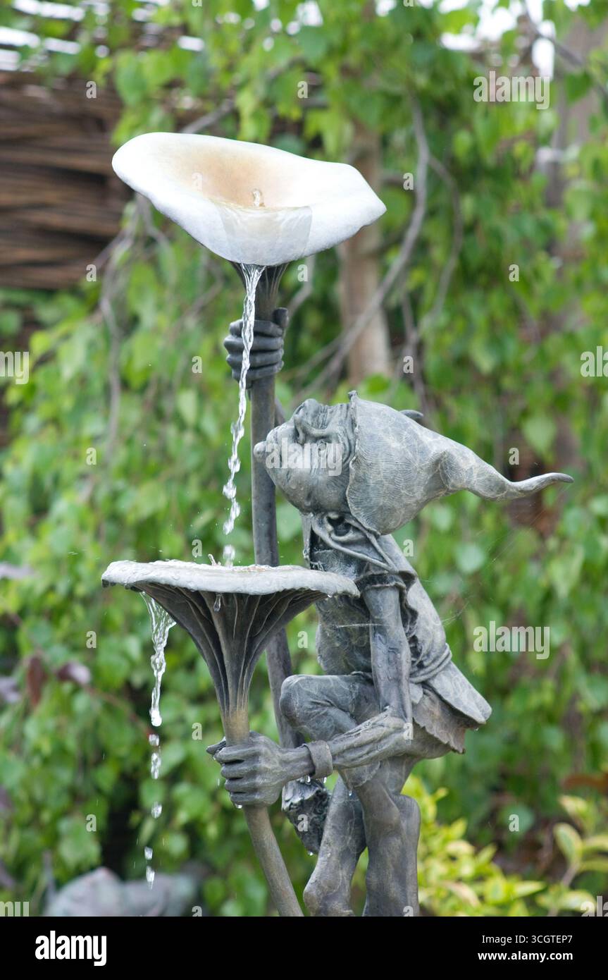 Un'affascinante statua raffigura un bambino che versa gioiosamente acqua da una fontana di fiori, circondato da lussureggianti piante verdi in un tranquillo giardino. Foto Stock