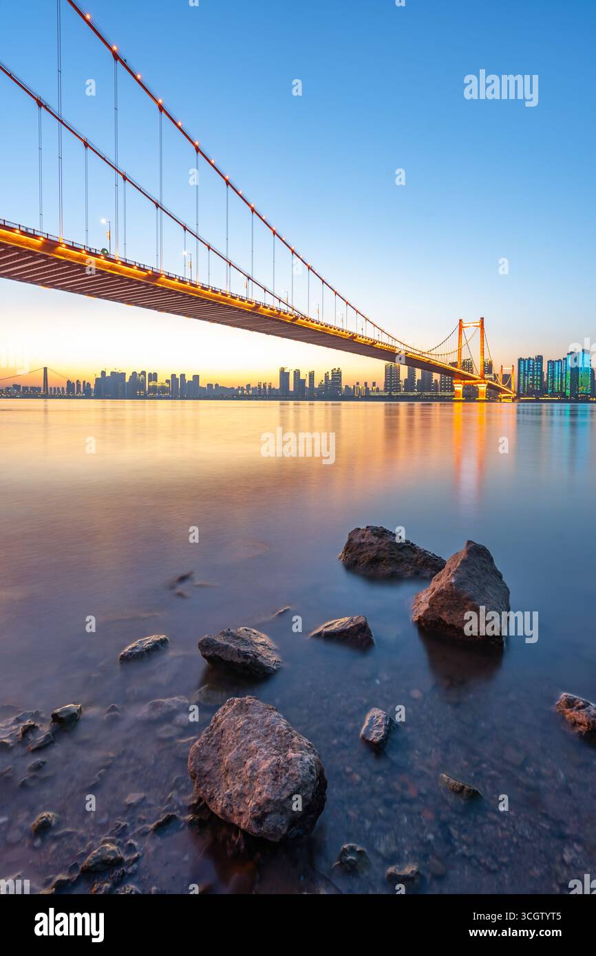 Il ponte sul fiume Yangtze di Wuhan si estende sull'acqua, illuminato dalle luci della città che si riflettono sulla tranquilla superficie sottostante. Le rocce emergono dal dolce f Foto Stock