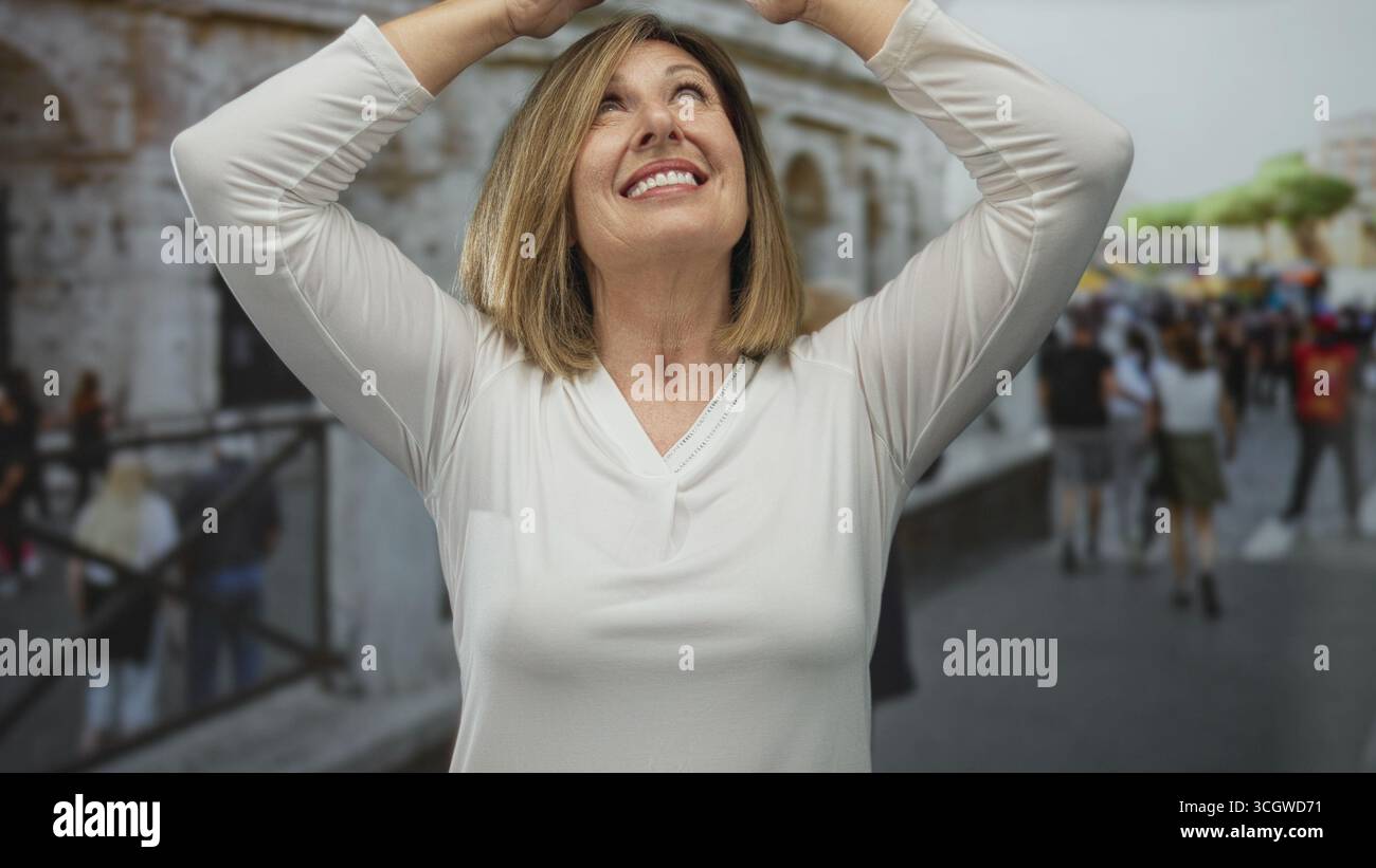 Donna caucasica di mezza età alza le mani sopra la testa sorridendo calorosamente in mezzo all'edificio del colosseo e alla folla offuscata di turisti; gioia. Foto Stock