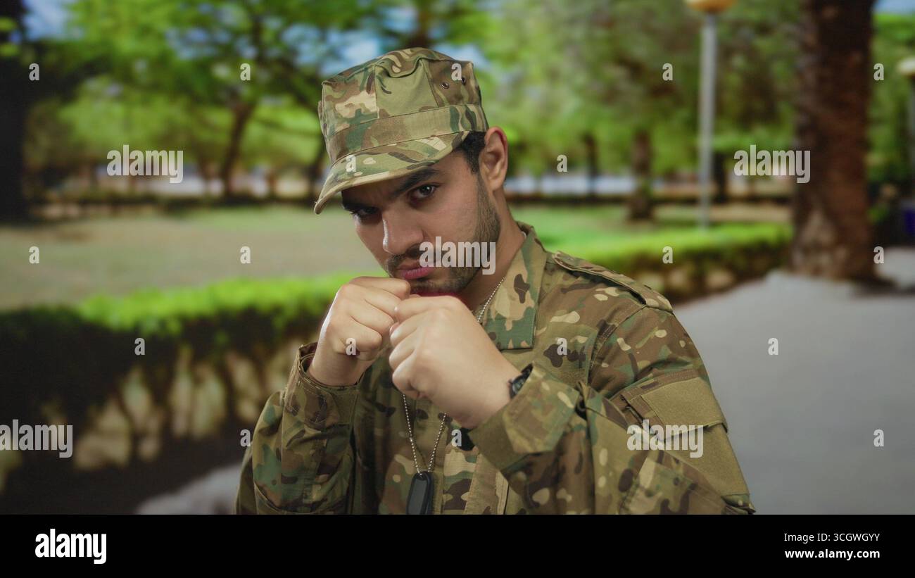 Uomo in uniforme mimetica solleva pugno armato in posa di combattimento in ambiente forestale con etichette per cani visibili; dovere coraggio. Foto Stock