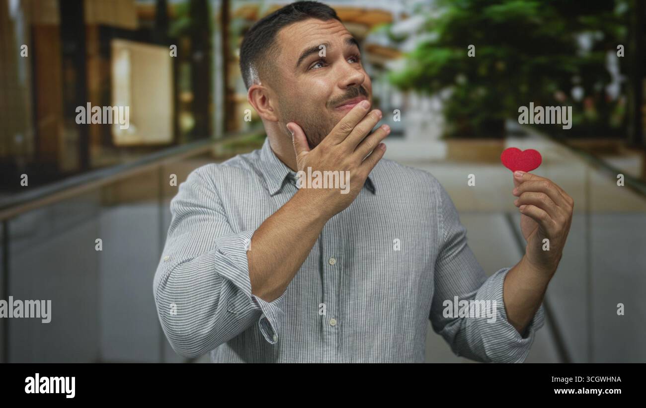 Il giovane ispanico tiene il cuore di carta rossa ritagliato con la mano sul mento per strada; riflesso romantico. Foto Stock