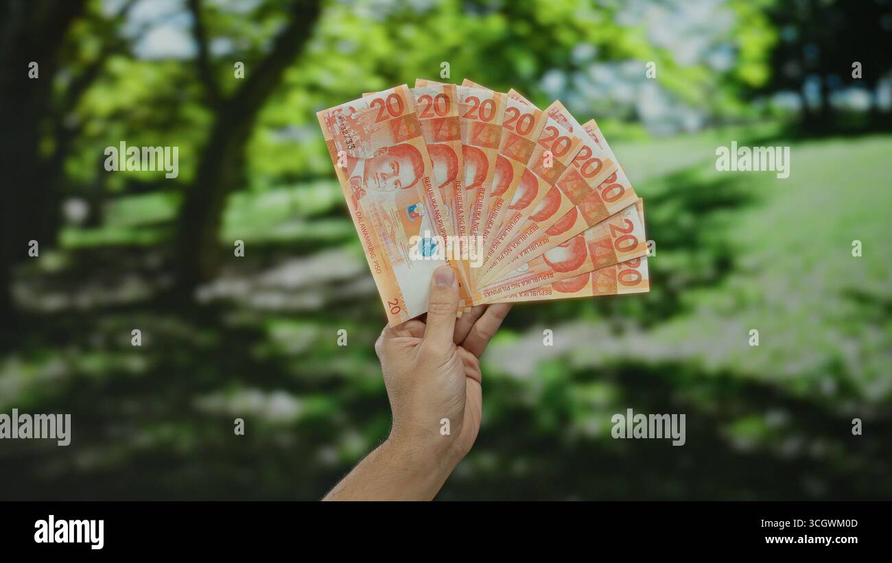 Uomo caucasico che tiene all'aperto diverse banconote in peso filippino in un parco, mettendo in evidenza un concetto di ricchezza o finanza in un ambiente naturale Foto Stock