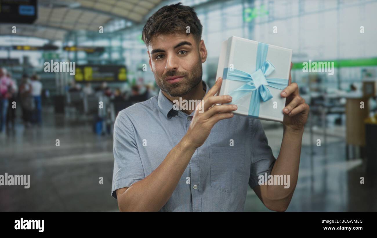 L'uomo tiene una confezione regalo avvolta vicino al viso nel terminal dell'aeroporto mentre sorride alla macchina fotografica; festeggia con gioia l'attesa. Foto Stock