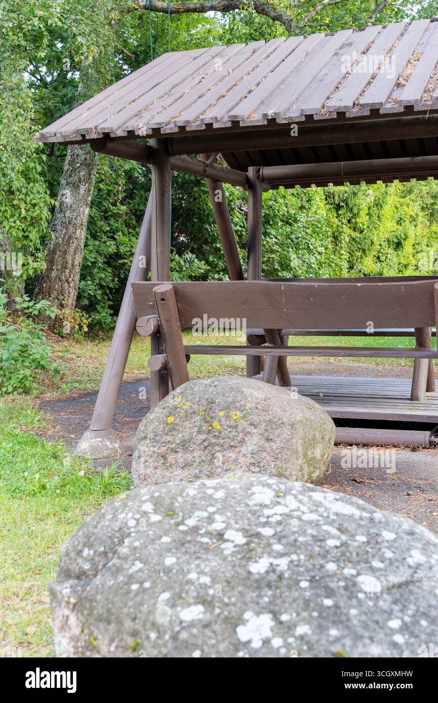 Una tranquilla panca in legno con tetto in un parco o giardino, circondata da lussureggianti alberi verdi e due grandi rocce in primo piano. Foto Stock