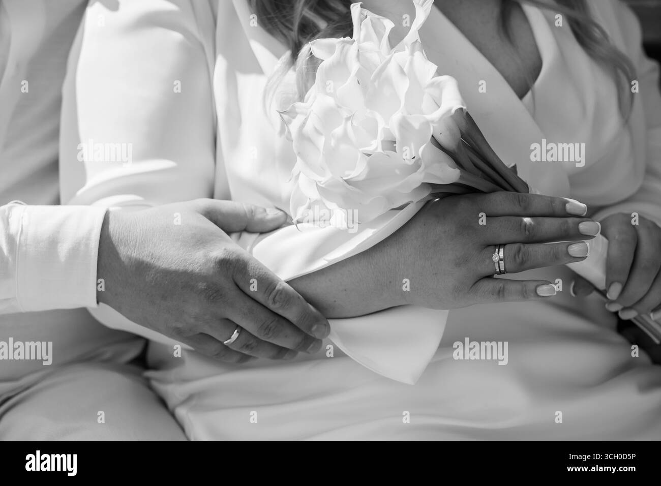 Un tenero primo piano delle mani di una coppia, delicatamente illuminato, che mostra le bande nuziali in argento mentre regge un mazzo di fiori bianchi, simboleggiando l'amore Foto Stock