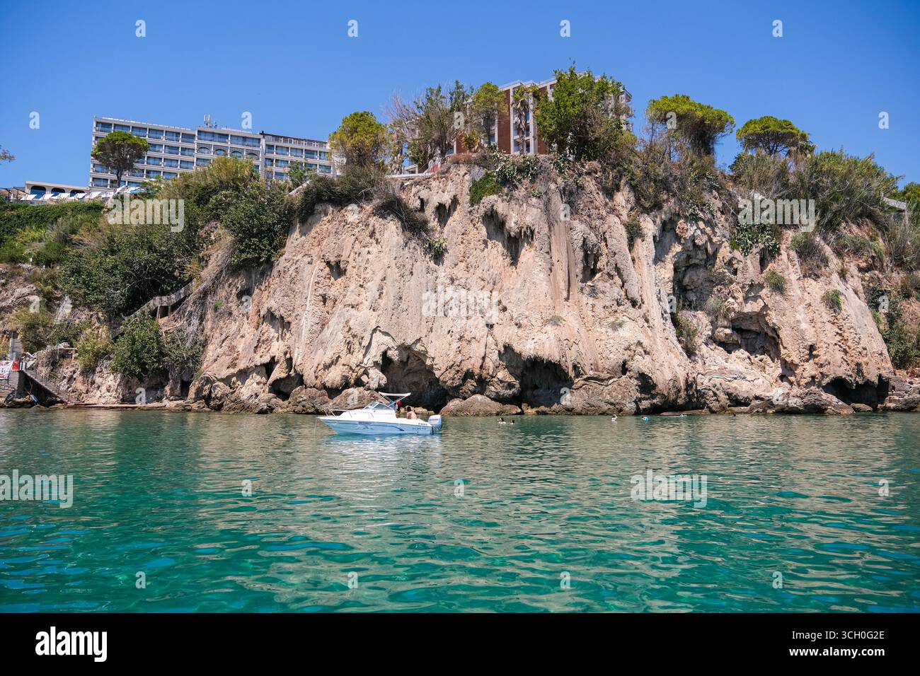 belek, Turkiye, 27.07.2025 piccola barca che galleggia su acque cristalline e verdi vicino a una scogliera rocciosa con un edificio alberghiero in cima sotto un cielo limpido. Foto Stock