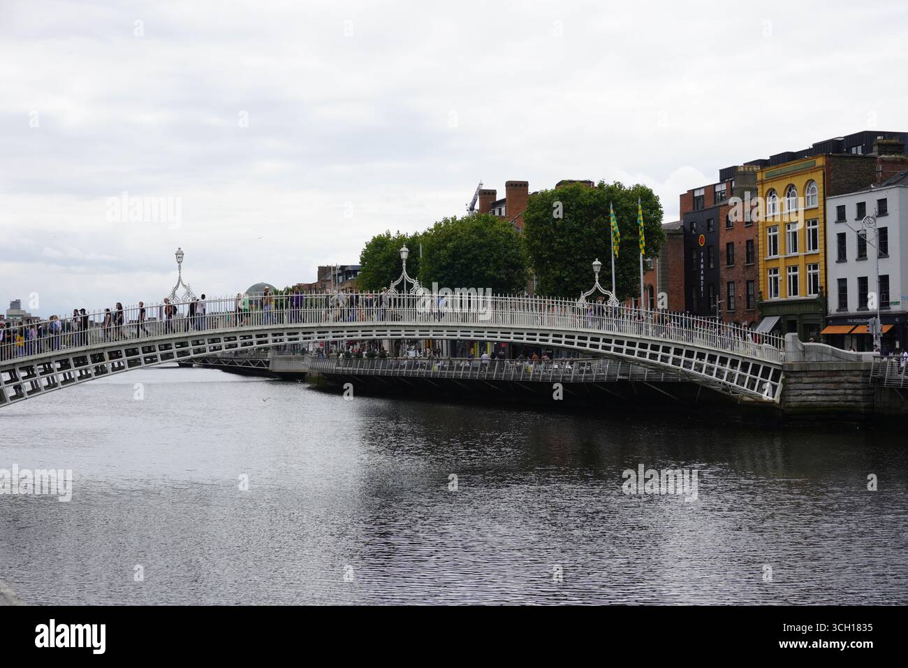 Un'immagine grandangolare del famoso Ha'penny Bridge di Dublino che attraversa il fiume Liffey, con persone che passeggiano e edifici storici che costeggiano le rive Foto Stock