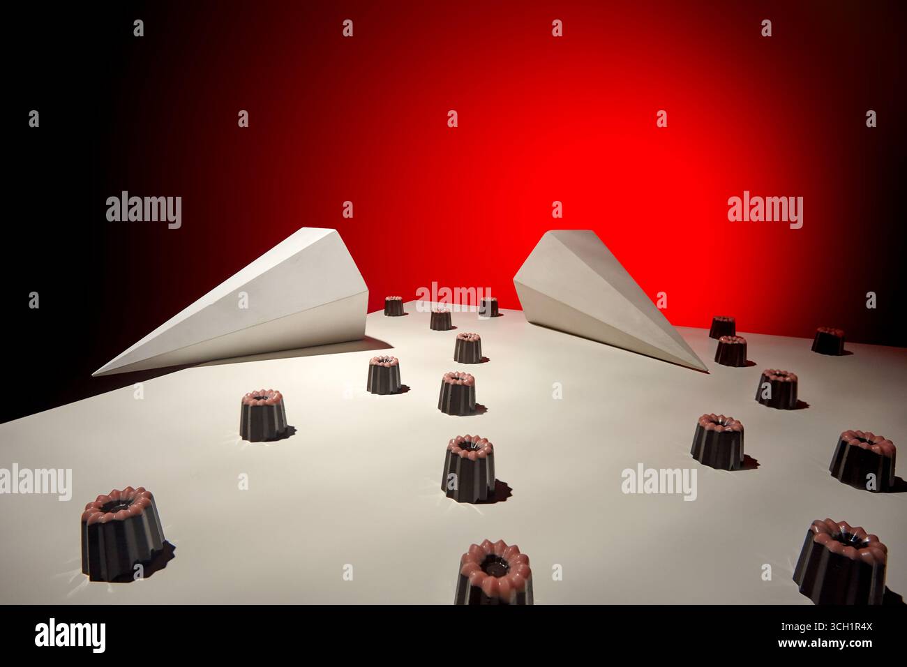 Una composizione concettuale di primo piano con caramelle di cioccolato disposte ordinatamente su una superficie bianca accanto a prismi geometrici astratti, evidenziati da una drammatica r Foto Stock
