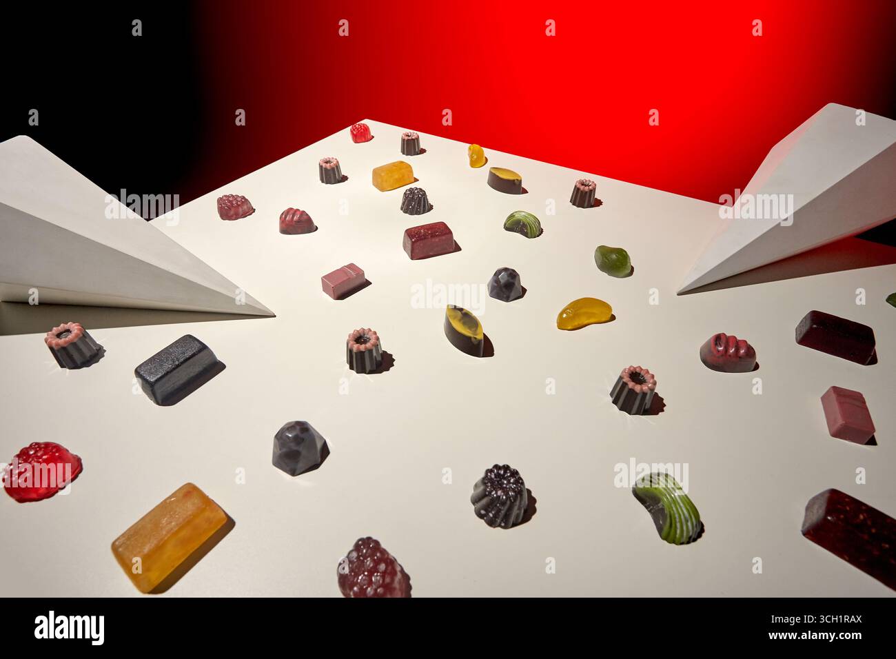 Una vivace natura morta che mostra caramelle colorate e assortite sparse su una superficie bianca con prismi astratti, evidenziati da audaci studio rosso e nero Foto Stock