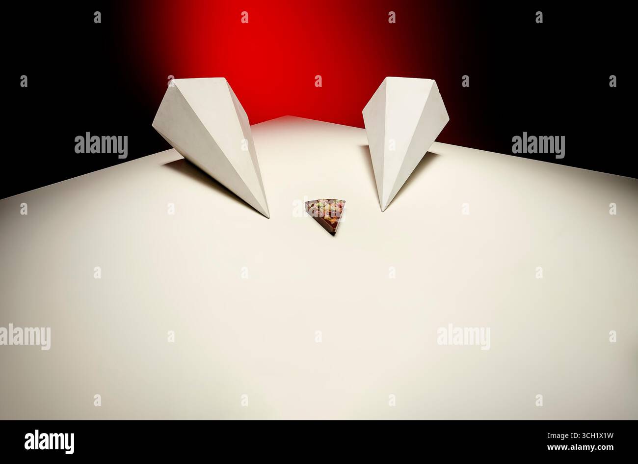 Una composizione minimalista con due strutture a prisma bianco angolari che incorniciano un singolo pezzo di barretta di cioccolato, adagiato drammaticamente su una b rossa e nera Foto Stock