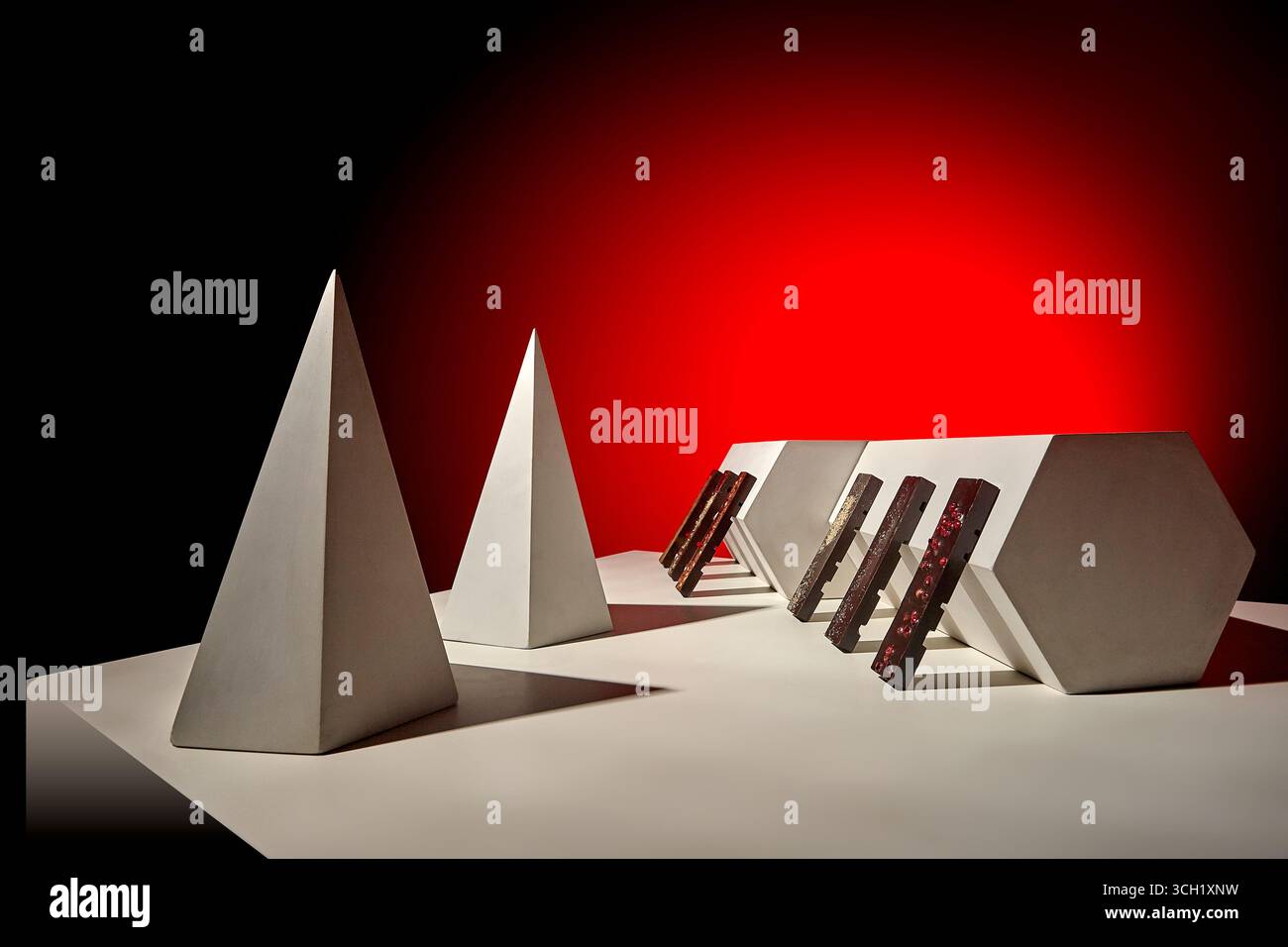 Una disposizione creativa di barrette di cioccolato poggiate su blocchi angolari accanto ad alte piramidi, fotografate su una superficie bianca con audace g rosso Foto Stock
