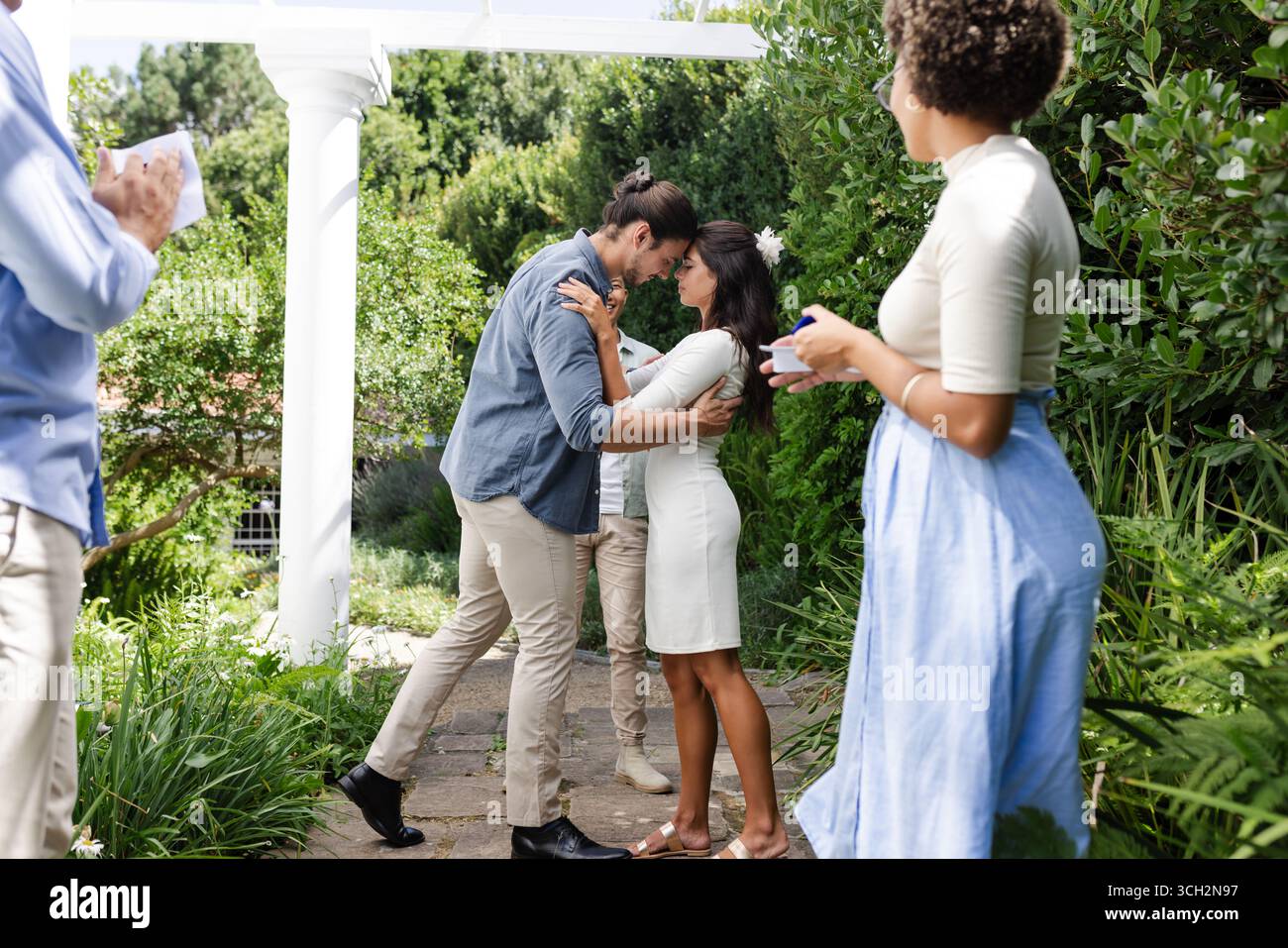 Coppia che abbraccia amorevolmente in giardino mentre gli amici festeggiano con gioia intorno a loro Foto Stock Coppia che abbraccia amorevolmente in giardino mentre gli amici festeggiano con gioia intorno a loro Foto Stock